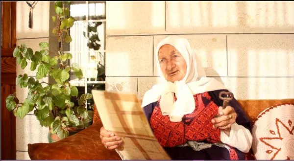 الحاجة فاطمة خلف (85 عامًا) ترفع كوشان لارض عائلتها في لفتا لمهجرة بيدها اليمنى ومفتاح بيتها المغتصب في الايسر